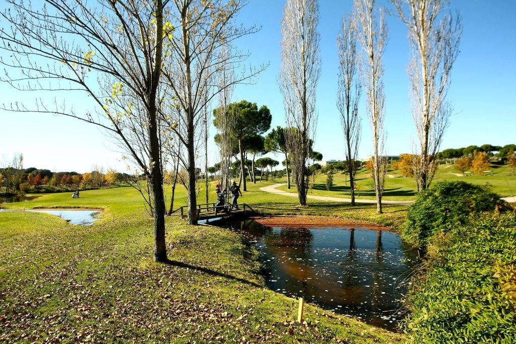 Autumn colours beside a fairway at Golf Santander in Madrid, with two golfers crossing a footbridge under a clear blue sky