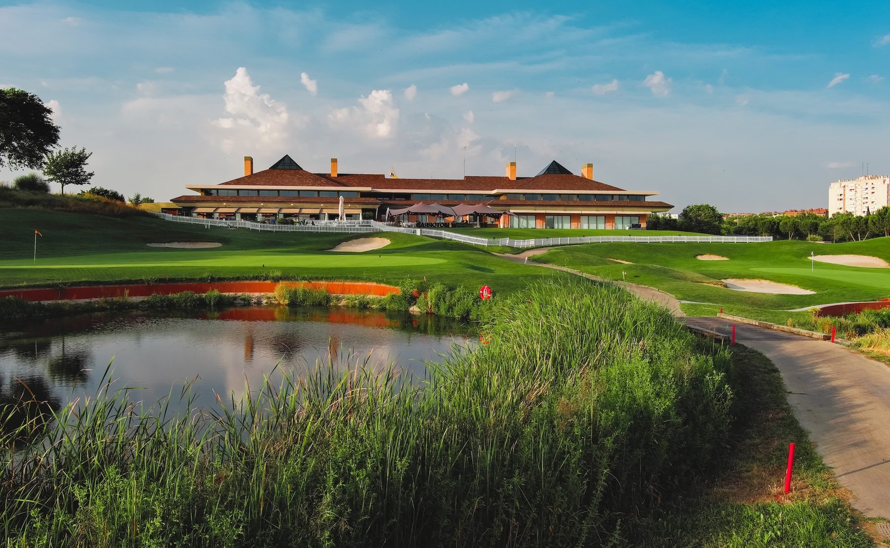 Clubhouse of Centro Nacional de Golf in Madrid seen across a pond and reeds, with a green and bunkers under a blue sky
