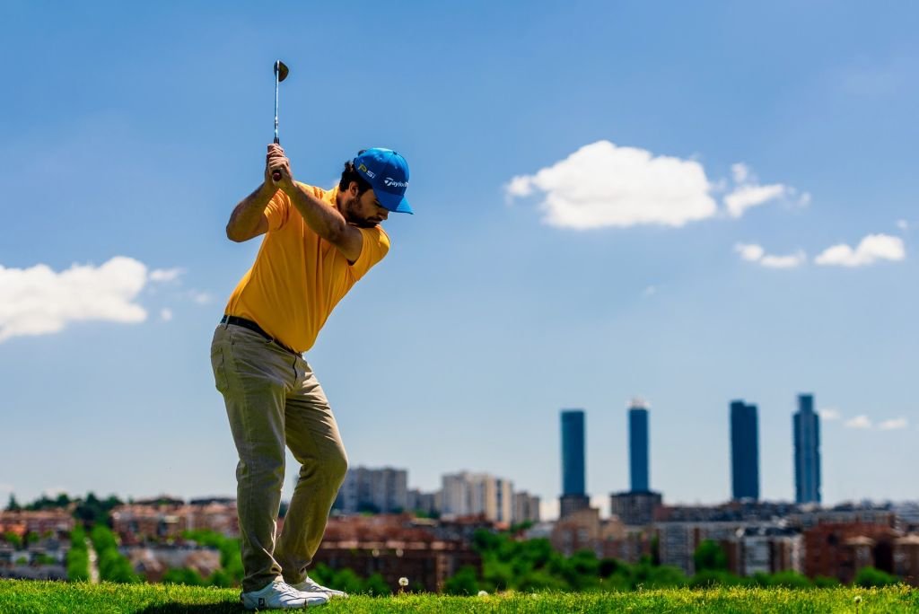 Golfer in a yellow polo mid-swing at Centro Nacional de Golf, with the Madrid skyline and the Cuatro Torres behind
